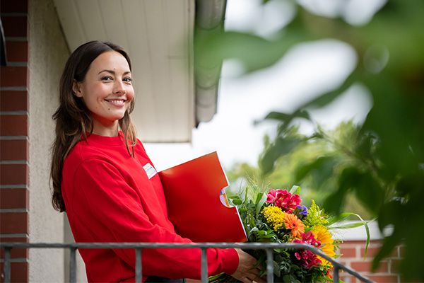 Smiling woman in a red sweater holds a bouquet of colorful flowers and a red folder on a balcony with greenery in the foreground.